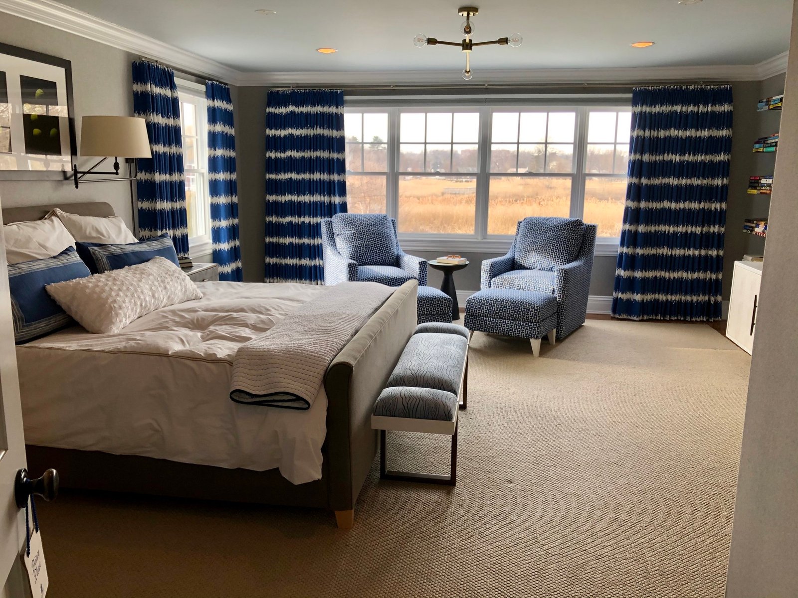 Primary suite seen from the doorway: an upholstered bed and bench with the sitting area and indigo drapery beyond