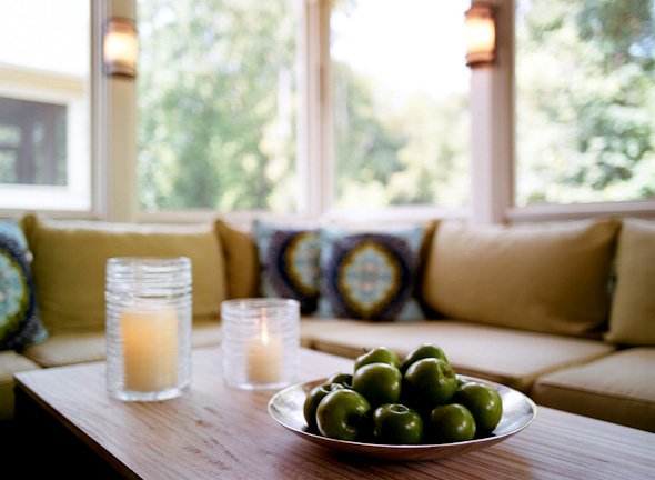 Hurricane candles and a bowl of green apples on the porch table before a sunlit sectional