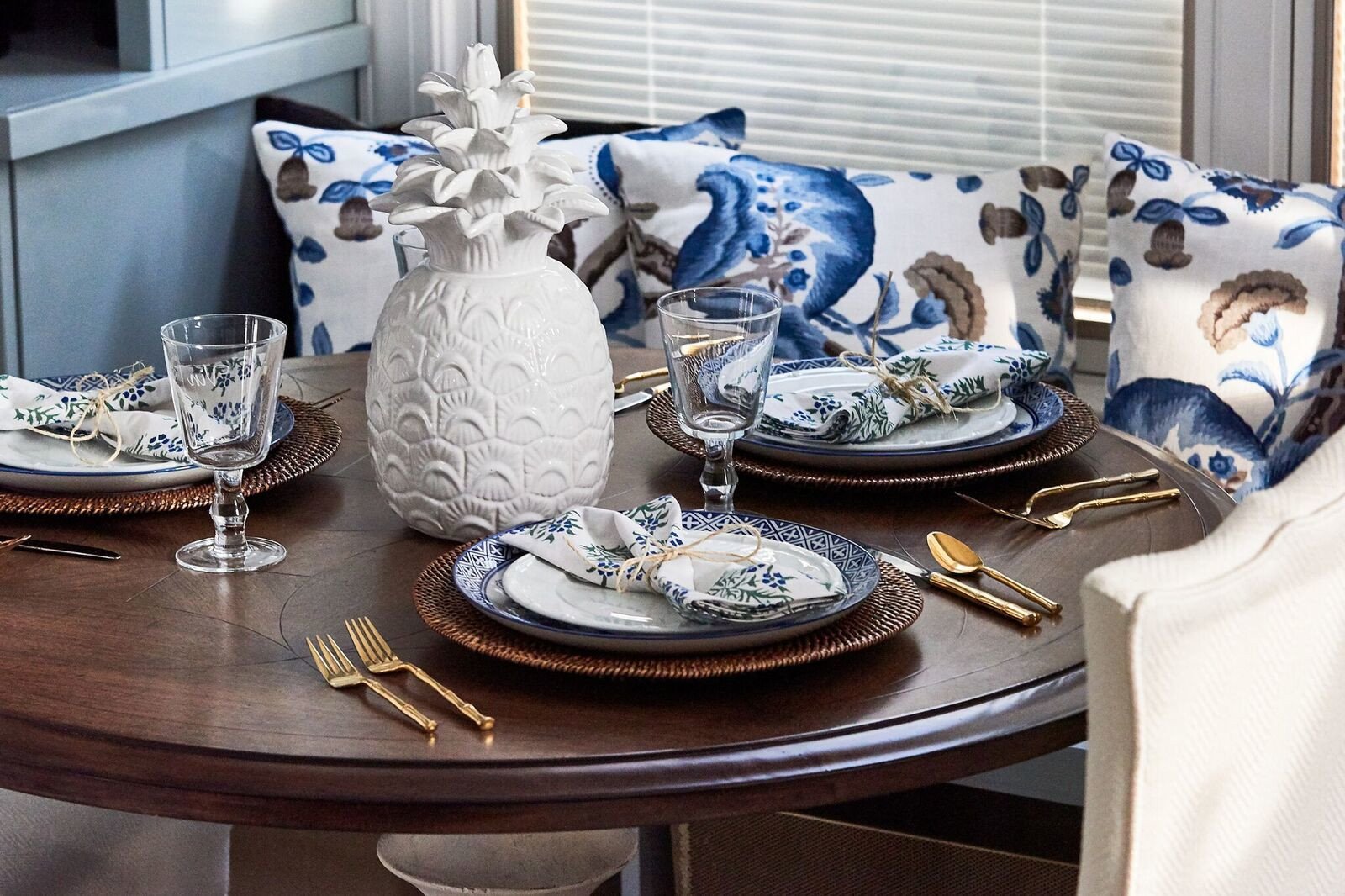 Round wood breakfast table set for guests with blue-and-white plates, floral napkins, clear goblets and gold flatware before an upholstered banquette