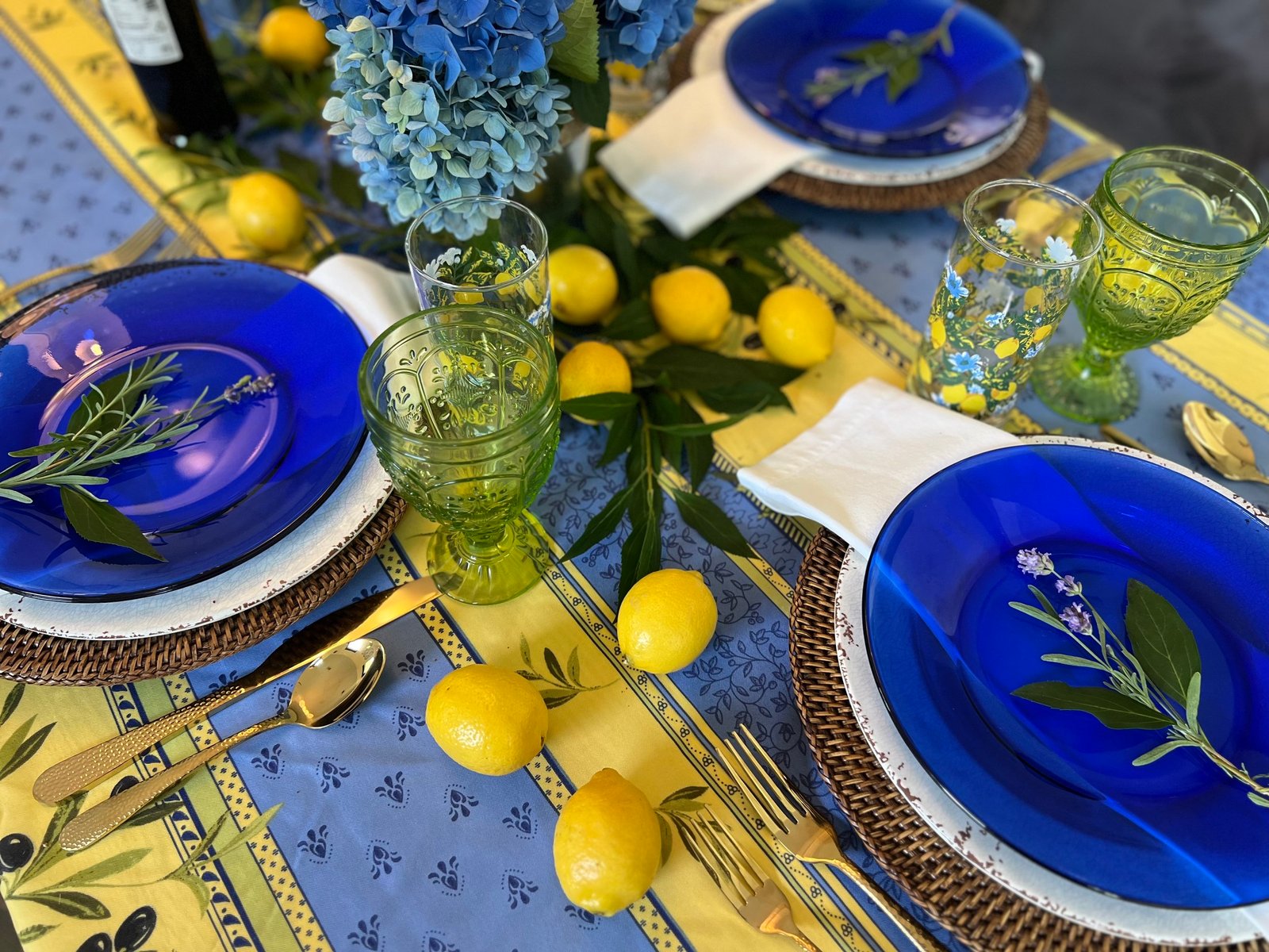 Tablescape of cobalt glass plates on rattan chargers, green pressed-glass goblets, lemons, blue hydrangeas and a Provençal yellow-and-blue tablecloth