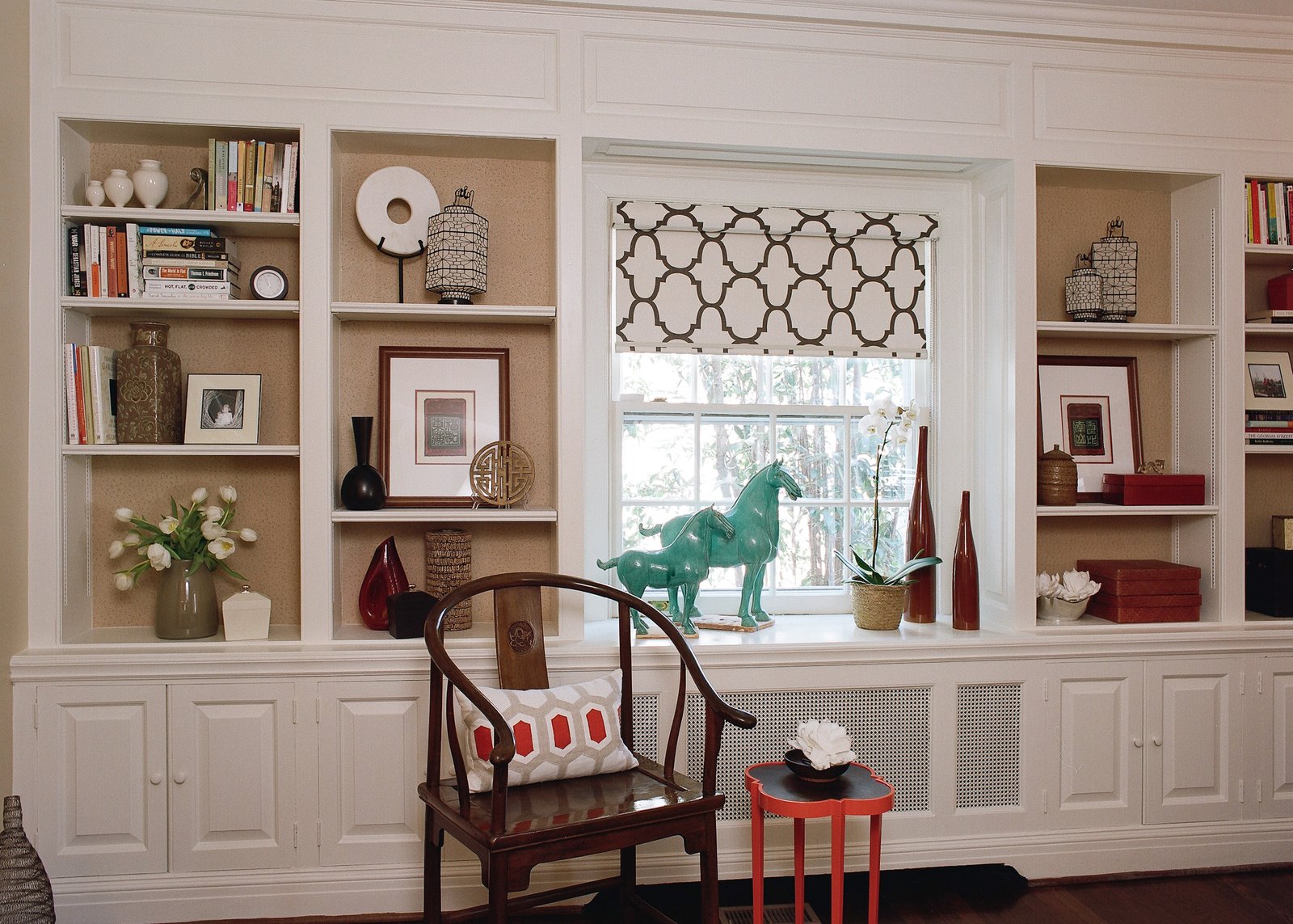 White built-in bookcases around a window with a geometric Roman shade, a horseshoe-back chair and red and turquoise accents