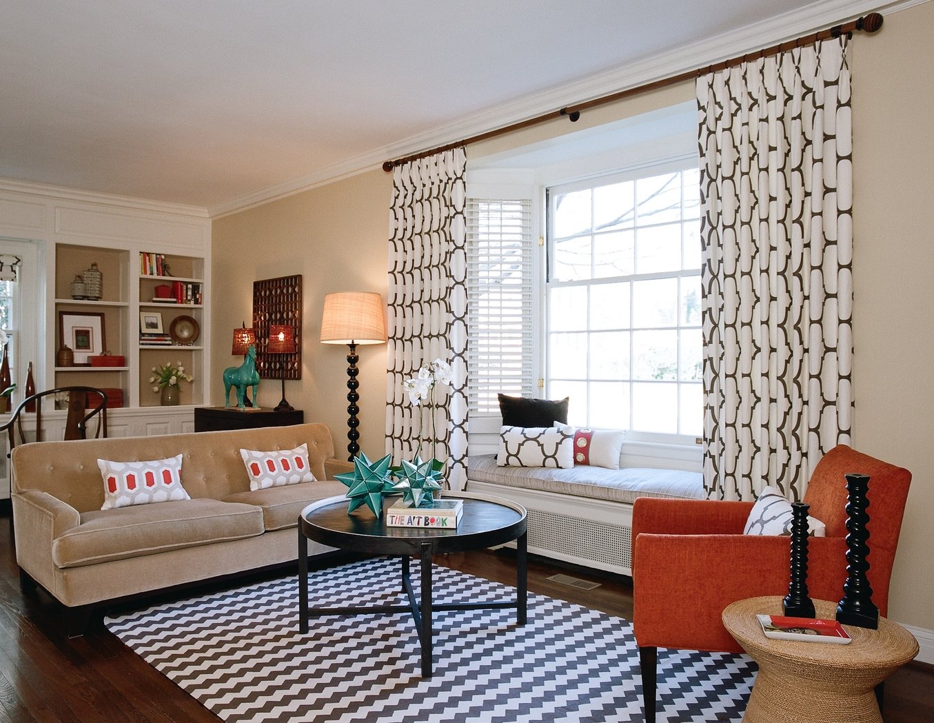 Living room with a camel sofa, chevron rug, black round cocktail table and a window seat framed by graphic drapery