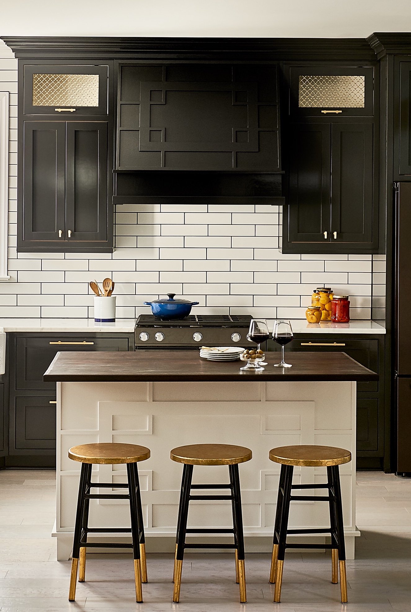 Black cabinetry and a paneled range hood over white subway tile, with a white paneled island, walnut top and three black and brass stools