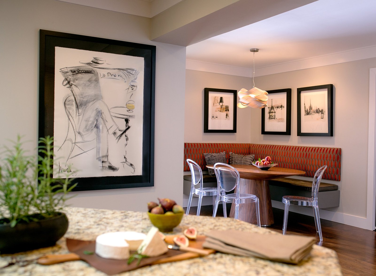 Cheese board on the island looking toward a breakfast banquette in rust upholstery with clear acrylic chairs and framed sketches
