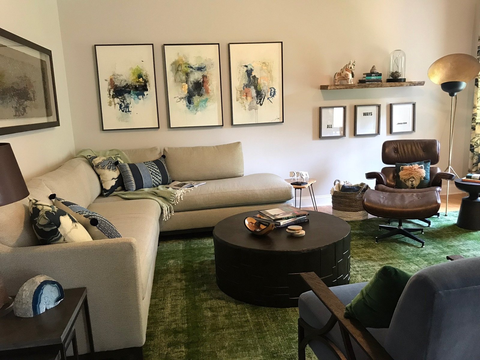 Living room with a green overdyed rug, a cream sectional, a leather lounge chair, a round black coffee table and three framed abstract watercolors