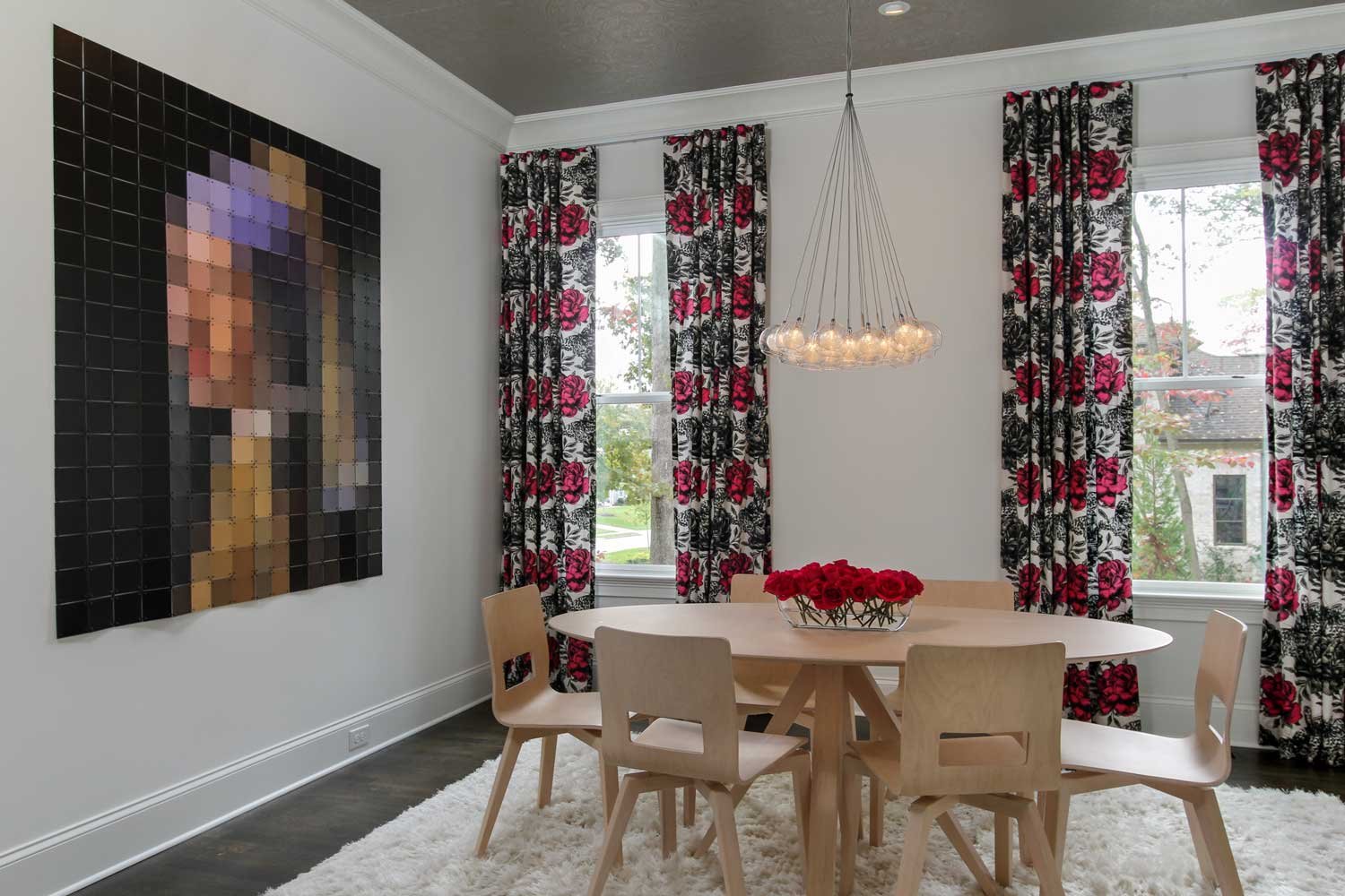 Dining room with a large pixel-mosaic portrait, a cascading glass-globe pendant, red-and-black floral draperies and a round wood table set with red roses