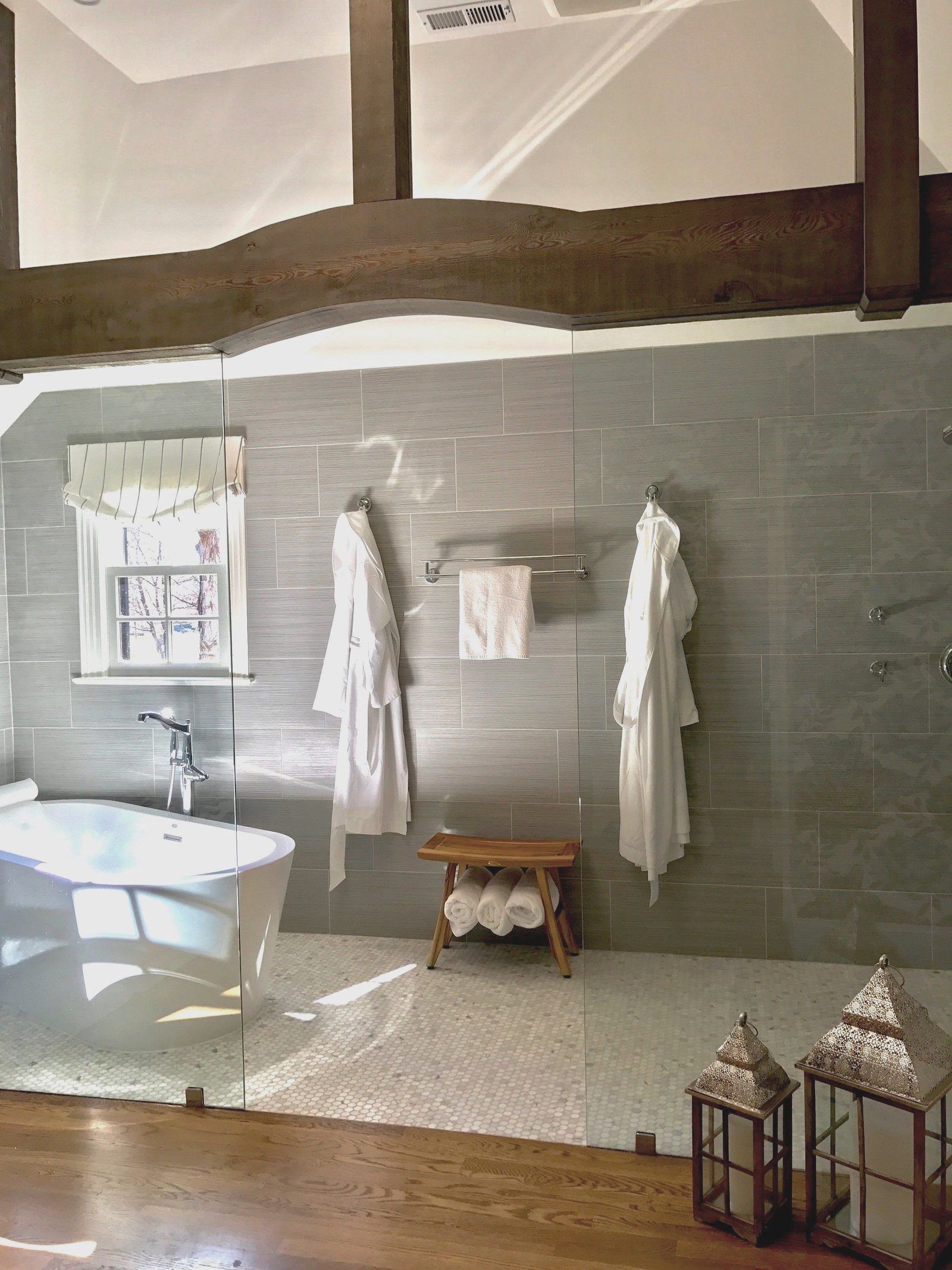 Primary bath beneath rustic timber beams with a freestanding soaking tub, an open glass shower in soft gray tile and white robes on hooks