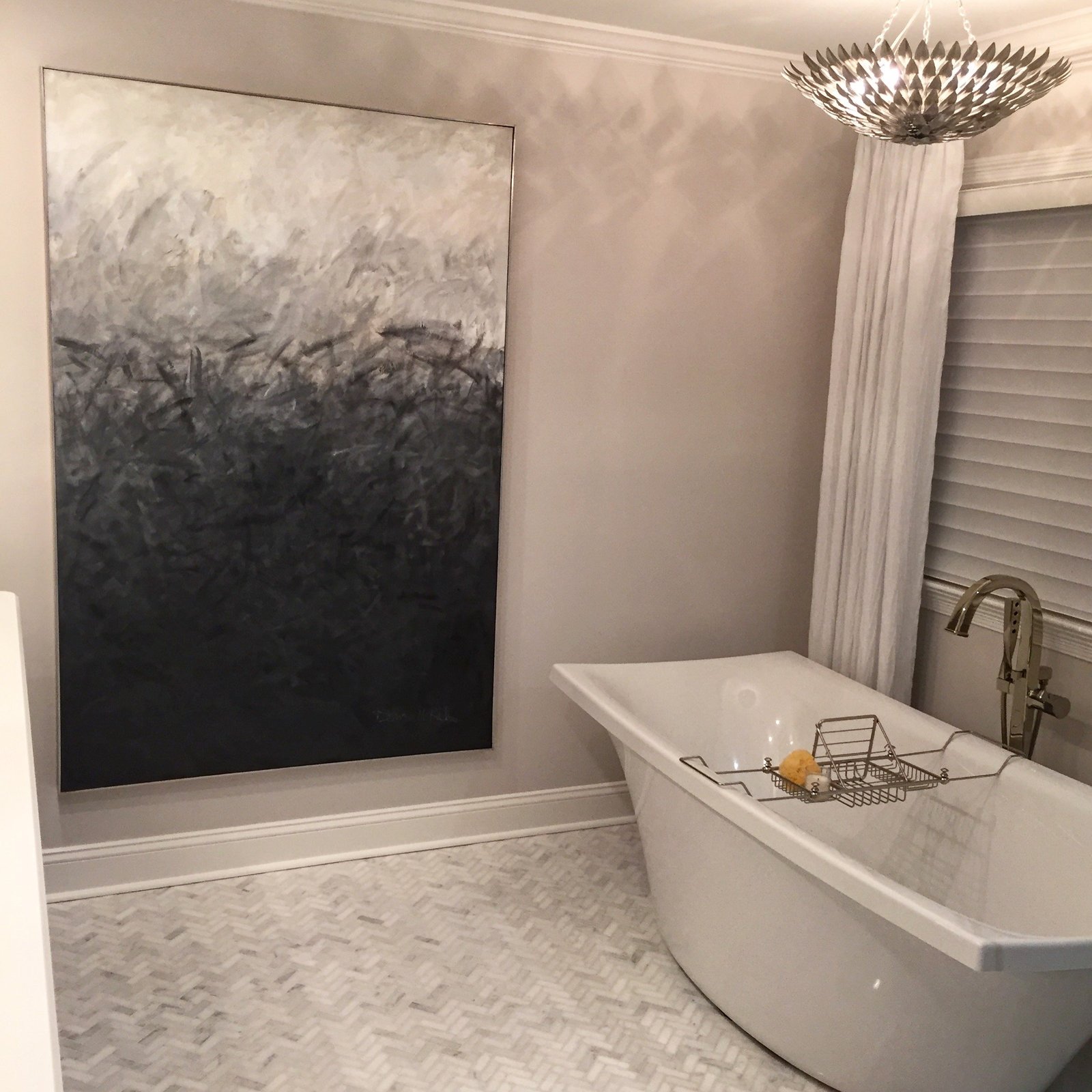 Freestanding tub beneath a large gray and black abstract painting, with a marble herringbone floor and a silver leaf ceiling fixture