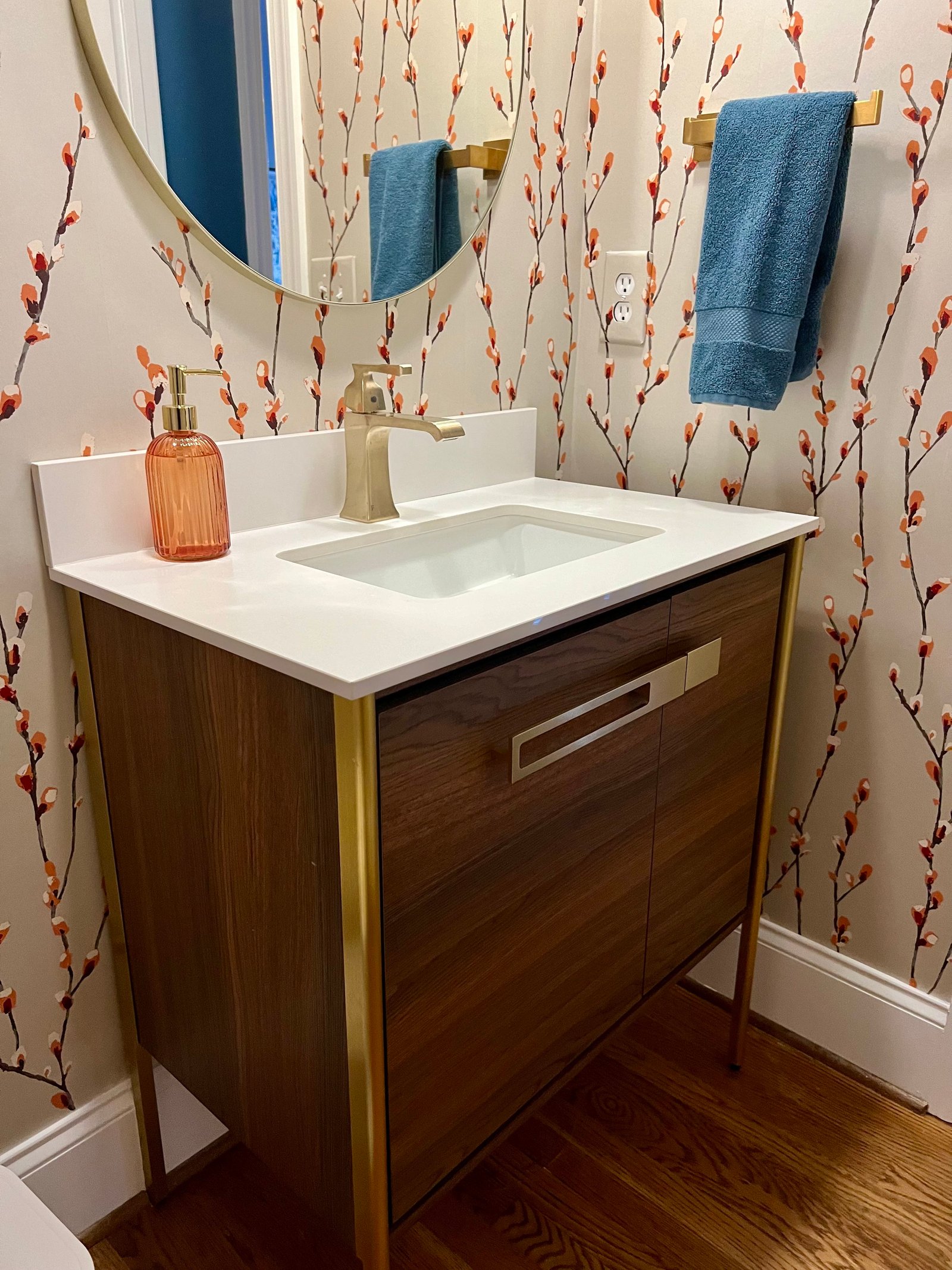 Powder room with blossom-branch wallpaper, a walnut and brass vanity, a round mirror and a blue hand towel