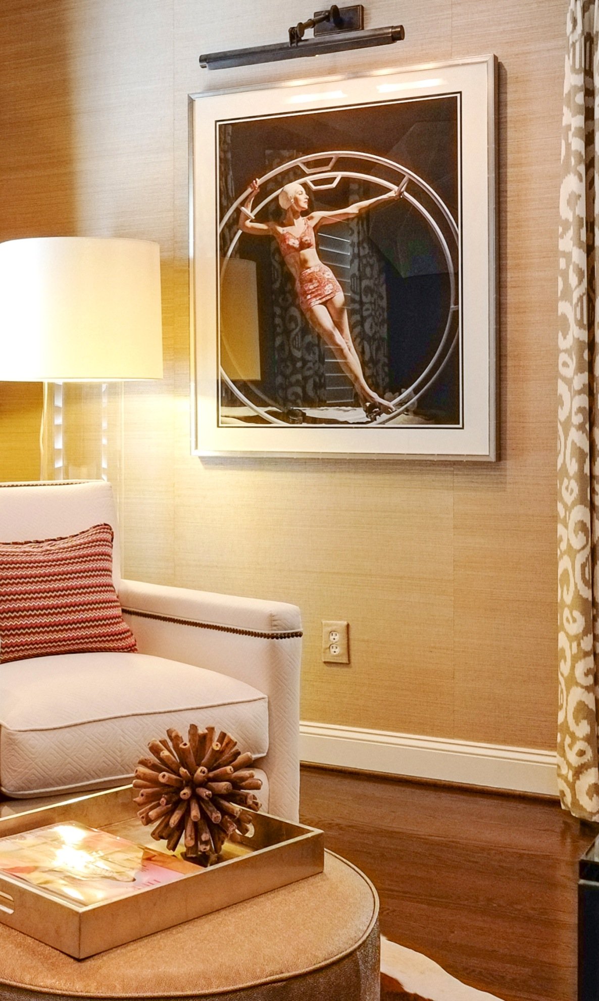 Reading corner with a cream nailhead armchair, a striped pillow, a lamp-lit framed photograph on grasscloth and a gilded tray on a round ottoman