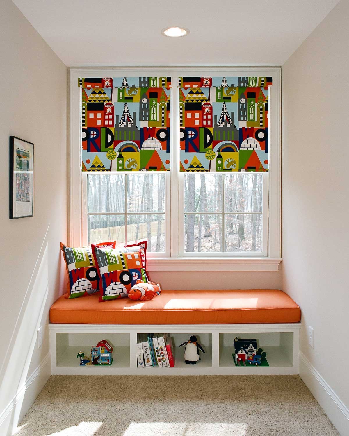 Child’s window seat with an orange cushion, graphic Roman shades and built-in book cubbies