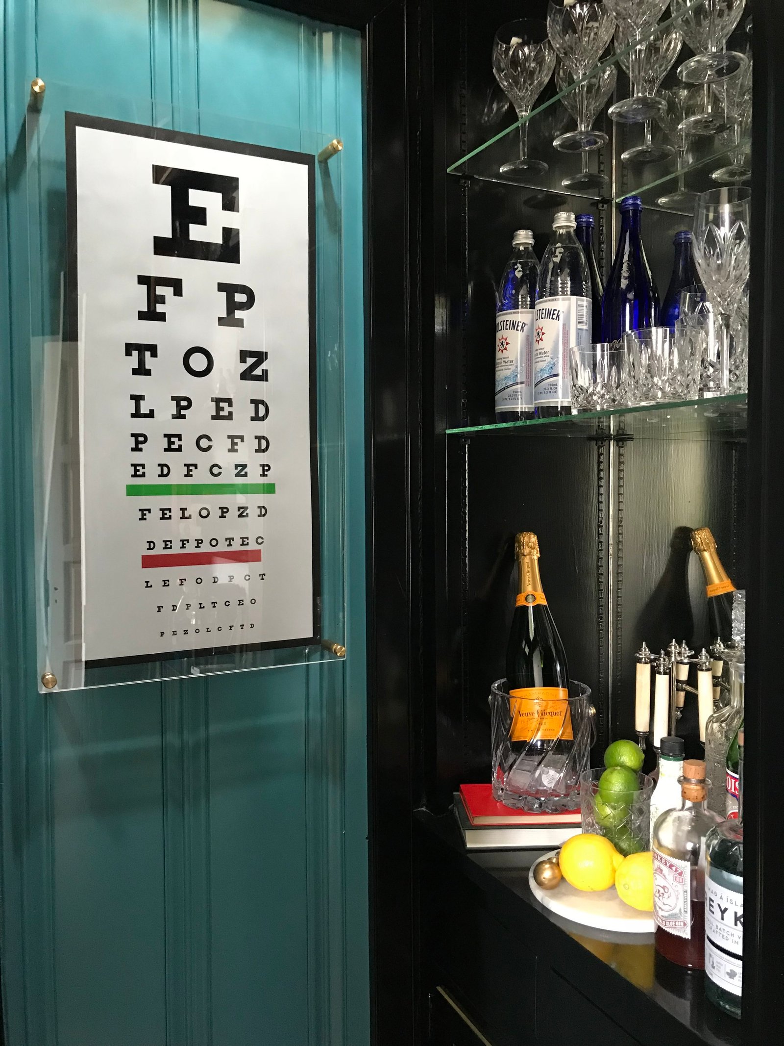 Built-in bar with teal lacquered doors, glass shelves of stemware, champagne on ice and an eye-chart artwork