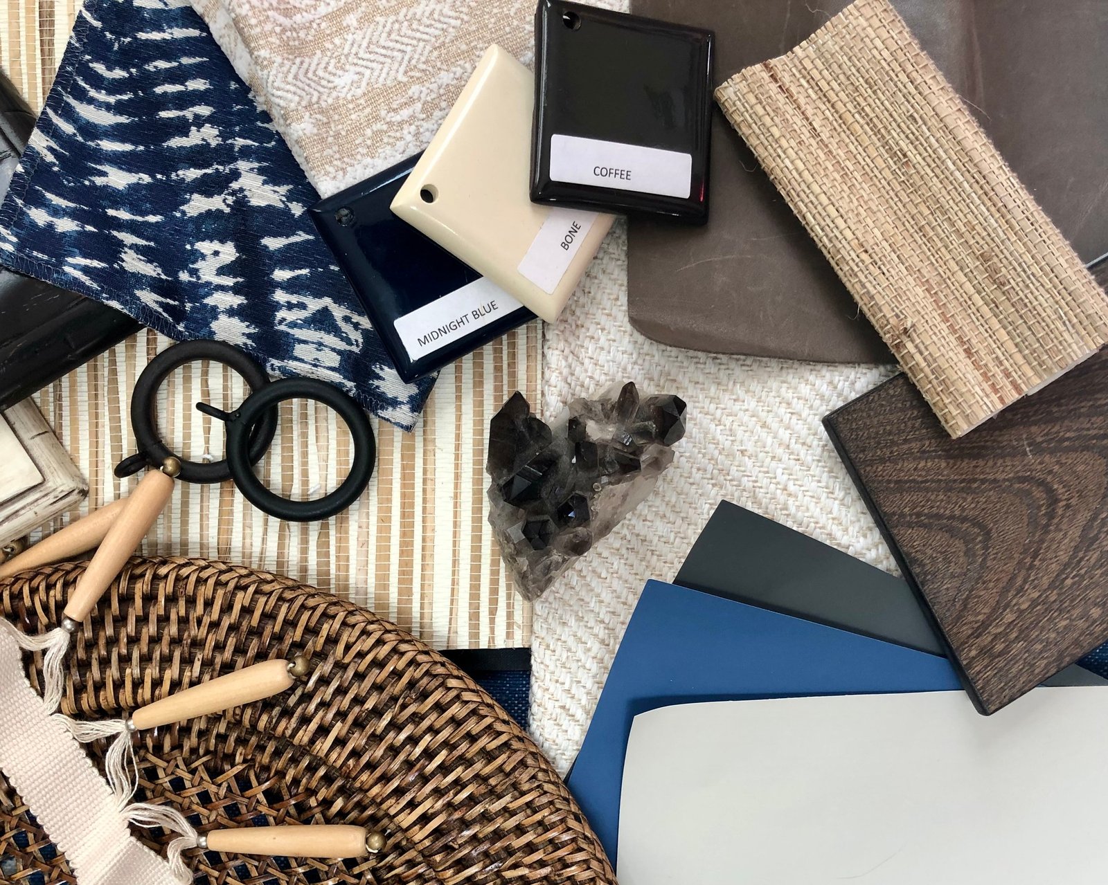 Studio flat-lay of navy ikat fabric, grasscloth and woven samples, lacquer finish chips in midnight blue, bone and coffee, a smoky quartz cluster and a rattan tray