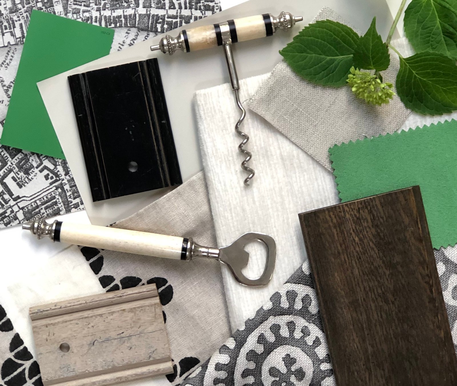 Flat-lay of green paint swatches, linen and map-print fabrics, ebonized and weathered moulding samples, a bone-handled corkscrew and bottle opener, and fresh hydrangea leaves